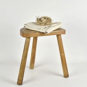 Peut inclure: Un petit tabouret en bois à trois pieds avec une assise ronde. Un tissu en lin blanc cassé plié et un petit nid d'oiseau avec des œufs reposent sur le tabouret. Le tout est sur fond blanc.