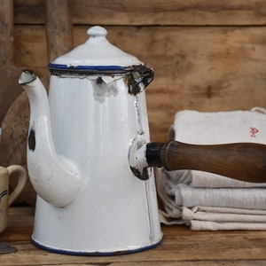 Peut inclure: Une cafetière ancienne en émail blanc avec une poignée en bois et un liseré bleu. Des traces d'usure témoignent de son âge. Une tasse avec des motifs floraux bleus et une cuillère sont en arrière-plan.