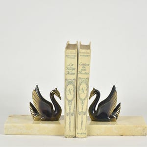 May include: A pair of swan bookends with a marble base, flanking two vintage books. The swans are black with gold-colored wings and beaks. The books are off-white with gold lettering and a decorative emblem.