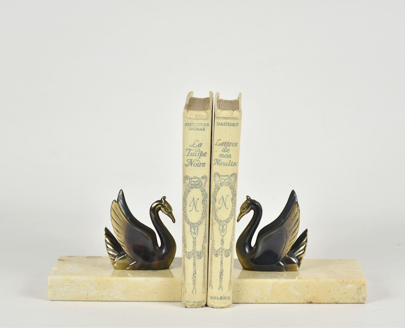 May include: A pair of swan bookends with a marble base, flanking two vintage books. The swans are black with gold-colored wings and beaks. The books are off-white with gold lettering and a decorative emblem.