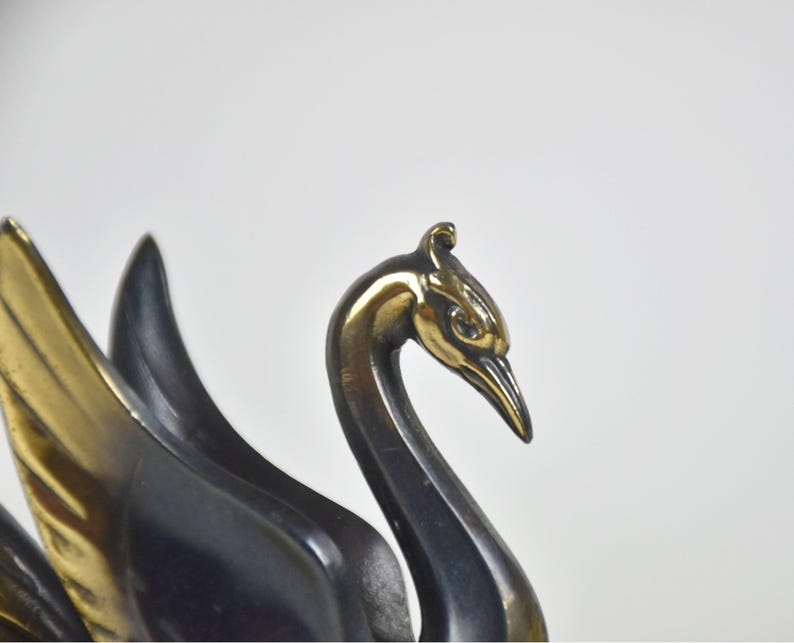 May include: A close-up of a bronze swan sculpture. The swan's head and beak are a shiny gold color, while the neck and body are a darker bronze. The wings are partially visible, with gold and dark bronze feathers.