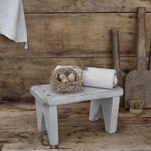 Puede incluir: Un pequeño taburete de madera azul claro desgastado sostiene un nido de pájaro con huevos moteados y un paño blanco enrollado. El fondo presenta tablones de madera y herramientas antiguas, creando una estética de granja.