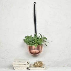 May include: A copper-coloured metal bowl planter with a rounded base, suspended by a long black strap. The planter contains a lush green sage plant. Below the planter sits a stack of folded off-white fabric cloths and a small woven bird nest.