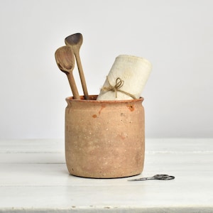 Op de afbeelding: Een cilindervormige terracotta pot met een gespikkeld, poreus oppervlak. In de pot staan twee donkerbruine houten lepels en een opgerold stuk gebroken wit textiel, vastgebonden met een dun touwtje. Een kleine metalen schaar ligt ernaast.