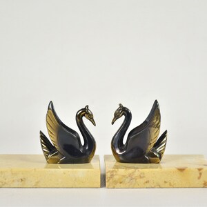 May include: A pair of swan figurines, each with a black body and gold-colored wings, perched on rectangular, beige marble bases. The swans face each other, creating a symmetrical display. Ideal for home decor.