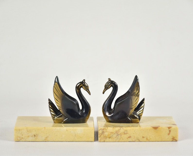 May include: A pair of swan figurines, each with a black body and gold-colored wings, perched on rectangular, beige marble bases. The swans face each other, creating a symmetrical display. Ideal for home decor.