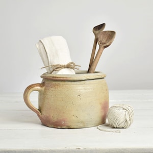 Op de afbeelding: Een beige keramieken pot met één handvat, gevuld met een gevouwen witte doek en twee houten lepels. Een kleine bol wit touw ligt naast de pot. Het keramische oppervlak heeft een gevlekte textuur met subtiele roodbruine kleurvariaties.