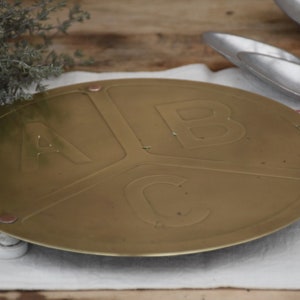 May include: A round, gold-colored metal tray with three sections. Each section has a raised letter, A, B, and C. The tray has three small white feet on the bottom.