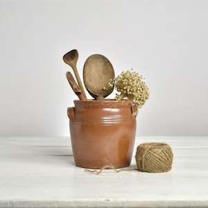 Peut inclure: Un pot en céramique brun avec des poignées, contenant des cuillères en bois et des fleurs séchées. Une pelote de ficelle est posée à côté. L'ensemble est disposé sur une surface blanche.