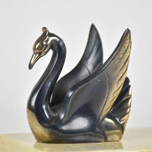 May include: A decorative swan figurine with a sleek, metallic finish. The swan's body is a dark, gunmetal gray, while the head and beak have a gold-toned accent. The wings are spread, and the sculpture sits on a light-colored base.