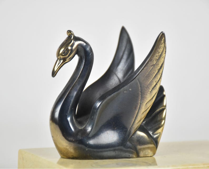 May include: A decorative swan figurine with a sleek, metallic finish. The swan's body is a dark, gunmetal gray, while the head and beak have a gold-toned accent. The wings are spread, and the sculpture sits on a light-colored base.
