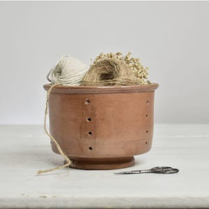 May include: A terracotta yarn bowl with a cylindrical shape and vertical rows of small holes along the side. Inside the bowl are two balls of yarn, one white and one brown, alongside dried flowers. A small pair of metal scissors lies nearby.