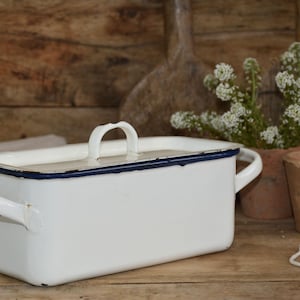 以下が含まれることがあります： 蓋と2つのハンドルが付いた白いエナメル製の長方形の容器。容器には濃い青色のトリムと蓋に白いハンドルが付いています。容器は木製の表面にあります。