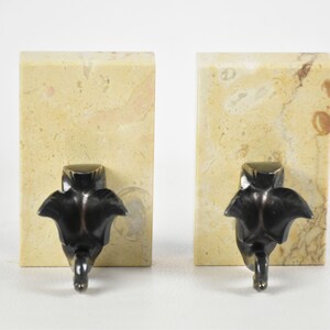 May include: Two bookends. Each bookend features a rectangular, beige marble base with brown veining. A dark bronze-colored, stylized leaf design is attached to the front of each base. The leaf design has a rounded top and a pointed bottom.