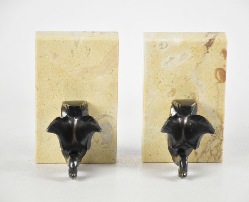 May include: Two bookends. Each bookend features a rectangular, beige marble base with brown veining. A dark bronze-colored, stylized leaf design is attached to the front of each base. The leaf design has a rounded top and a pointed bottom.