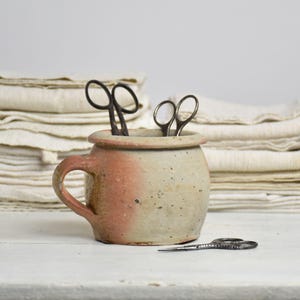 Puede incluir: Un pequeño recipiente de cerámica redondo y moteado con una sola asa, que presenta una mezcla de tonos beige y terracota. Dos pares de tijeras de metal oscuro están colocados dentro del recipiente y otro par de tijeras descansa en la superficie.
