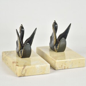 May include: A pair of swan bookends. Each bookend features a black swan sculpture with gold accents, mounted on a rectangular, cream-colored marble base. The swans are depicted with their wings spread. The bookends are designed for decorative and functional use.