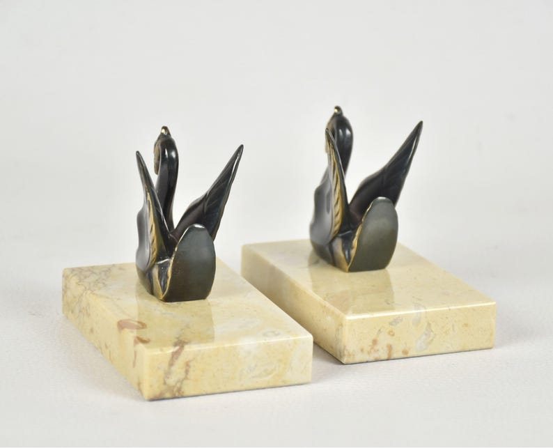 May include: A pair of swan bookends. Each bookend features a black swan sculpture with gold accents, mounted on a rectangular, cream-colored marble base. The swans are depicted with their wings spread. The bookends are designed for decorative and functional use.