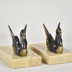 May include: Two swan figurines with outstretched wings, crafted from dark metal with gold accents, are mounted on rectangular, cream-colored marble bases. The swans face opposite directions, creating a balanced composition. The background is plain white.