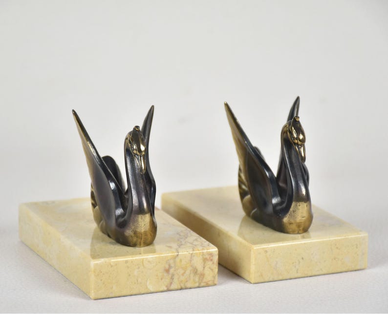 May include: Two swan figurines with outstretched wings, crafted from dark metal with gold accents, are mounted on rectangular, cream-colored marble bases. The swans face opposite directions, creating a balanced composition. The background is plain white.