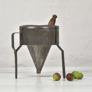 Könnte beinhalten: Eine vintage Passiermühle aus Metall mit einem konischen, perforierten Sieb, einer dunkelgrauen Oberfläche und einem Holzstößel. Das Küchengerät steht auf drei Metallbeinen, hat einen seitlichen Griff und wird von vier kleinen Feigen begleitet.
