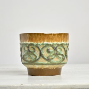 May include: A ceramic bowl featuring a brown glazed rim and base with a central band of raised, textured green and dark brown scrollwork patterns. The vessel has a wide, open top and a tapered base, showcasing a multi-toned, earthy color palette.