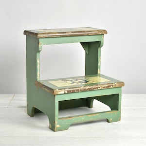 Peut inclure: Un marchepied en bois à deux niveaux peint en vert sauge atténué avec des bords visiblement usés. Les marches supérieure et inférieure présentent un panneau rectangulaire incrusté avec un motif de coq. La finition montre des signes d'usure.