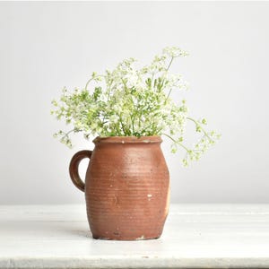 Puede incluir: Un jarrón de cerámica marrón rústico con asa, lleno de delicadas flores blancas y follaje verde. El jarrón tiene una superficie texturizada y una forma ligeramente irregular, lo que sugiere que está hecho a mano. Las flores añaden un toque de frescura.