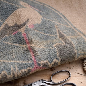 Peut inclure: Gros plan d'un sac en toile de jute bleu et brun avec un motif de cigogne blanche. Le sac est plié et le motif est partiellement masqué. Le sac est posé sur une surface en bois avec une paire de ciseaux et une bobine de ficelle.