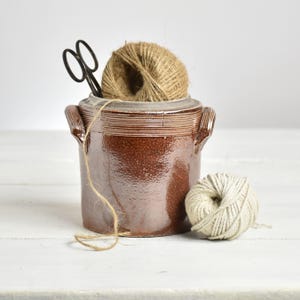 Peut inclure: Un pot en céramique marron avec des poignées, contenant une pelote de ficelle et une paire de ciseaux noirs. Une deuxième pelote de fil est posée à côté du pot. Le pot a une surface texturée et un bord décoratif.