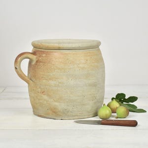 Op de afbeelding: Een cilindrische stenen pot met één handvat aan de zijkant en een bijpassend deksel. De pot heeft een gevlekte beige en bruine afwerking met een zichtbare kleitextuur. Naast de pot liggen drie kleine groene vijgen en een klein mesje.