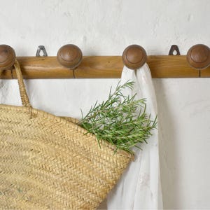 Może przedstawiać: Drewniany wieszak na ubrania z czterema zaokrąglonymi gałkami. Z jednego guzika wisi tkana słomiana torba, a z innych zwisają biała szmatka i gałązki rozmarynu. Wieszak jest zamontowany na białej ścianie.