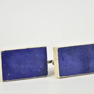 May include: A pair of rectangular cufflinks with a textured, deep blue surface and off-white borders. The cufflinks have a silver-colored metal backing.