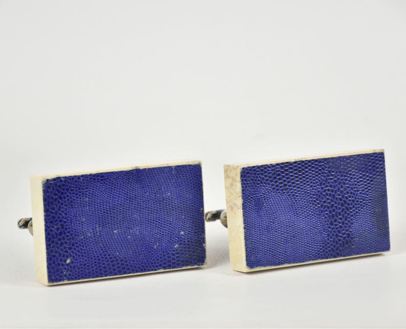 May include: A pair of rectangular cufflinks with a textured, deep blue surface and off-white borders. The cufflinks have a silver-colored metal backing.