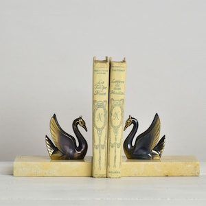 Pair of vintage Art Deco swan bookends made of dark patinated metal with gold highlights. The graceful swan figures sit on rectangular cream-colored marble bases. Two old yellowed French books by Dumas and Daudet are held between them on a white surface. Captures a sophisticated French Country Decor library aesthetic.