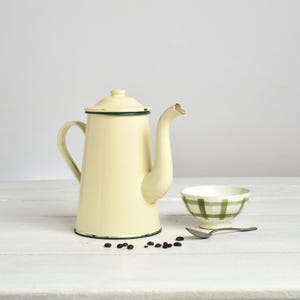 Peut inclure: Une cafetière en émail crème avec un couvercle et un bec courbé. Un petit bol à carreaux verts et blancs et une cuillère sont à côté de la cafetière. Des grains de café sont éparpillés sur la surface blanche.