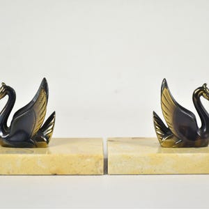May include: A pair of swan bookends. Each bookend features a black swan with gold-colored accents on a rectangular, cream-colored base. The swans have their wings spread and their heads held high, creating an elegant and decorative item.