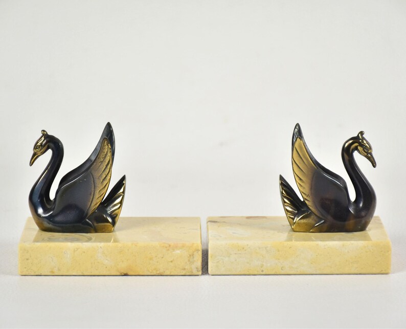 May include: A pair of swan bookends. Each bookend features a black swan with gold-colored accents on a rectangular, cream-colored base. The swans have their wings spread and their heads held high, creating an elegant and decorative item.
