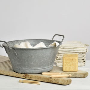 May include: A vintage metal washtub with handles, containing white fabric, rests on a wooden washboard. Wooden pegs and a bar of Marseille soap are nearby. A stack of folded linen towels is in the background.