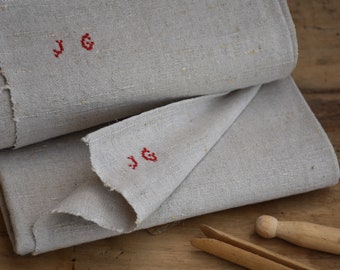 May include: Two pieces of gray linen fabric folded together. The top piece has the initials "JG" embroidered in red thread. The bottom piece has the same initials embroidered in red thread.