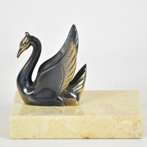 May include: A bronze-toned swan figurine with a gold beak and wing accents, perched on a rectangular marble base. The swan's feathers are detailed, and the sculpture has a vintage aesthetic. The base is a light beige color.
