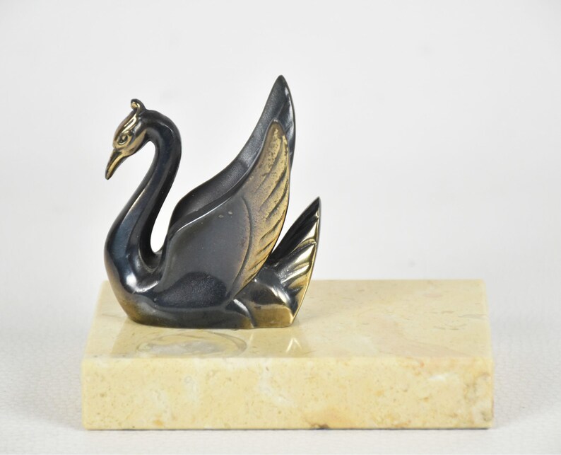 May include: A bronze-toned swan figurine with a gold beak and wing accents, perched on a rectangular marble base. The swan's feathers are detailed, and the sculpture has a vintage aesthetic. The base is a light beige color.