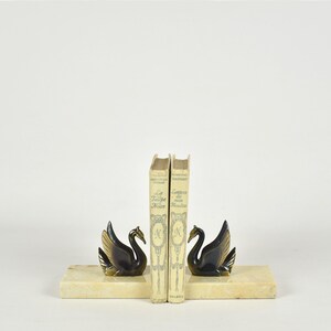 May include: A pair of swan bookends with gold-toned wings and black bodies, resting on rectangular, cream-colored bases. Two vintage books stand between the bookends. The bookends add a decorative touch to any bookshelf.