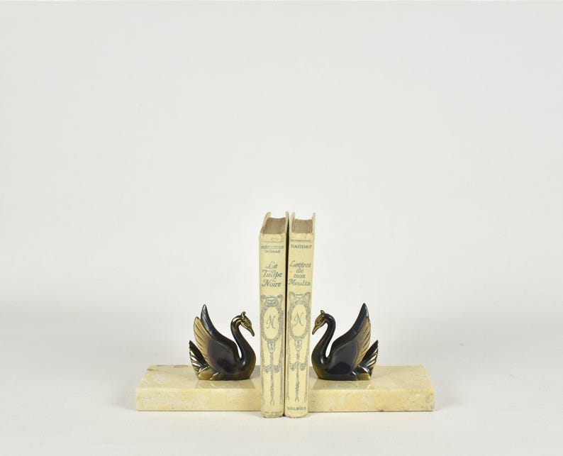 May include: A pair of swan bookends with gold-toned wings and black bodies, resting on rectangular, cream-colored bases. Two vintage books stand between the bookends. The bookends add a decorative touch to any bookshelf.
