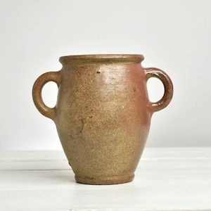 Può includere: Un recipiente rustico in terracotta con corpo arrotondato, bocca larga e due manici laterali simmetrici. La superficie presenta una smaltatura maculata marrone e marrone rossiccio con un aspetto strutturato, usurato e imperfezioni visibili.