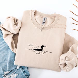 Puede incluir: Sudadera beige con el texto "Stupid Canadian Wolf bird" y un gr&aacute;fico de un somorgujo. La sudadera est&aacute; doblada, con un collar plateado y una etiqueta visible. El fondo incluye unos vaqueros azules y ramitas decorativas.