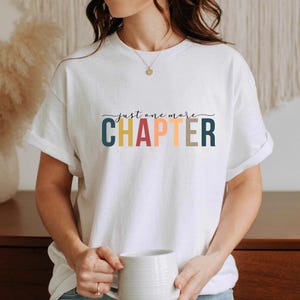 Könnte beinhalten: Weißes T-Shirt mit dem Text "just one more CHAPTER" in einer verspielten Schriftart. Das Wort "CHAPTER" ist in Blau, Gelb, Orange und Braun gehalten. Die Person, die das T-Shirt trägt, hält eine weiße Tasse.