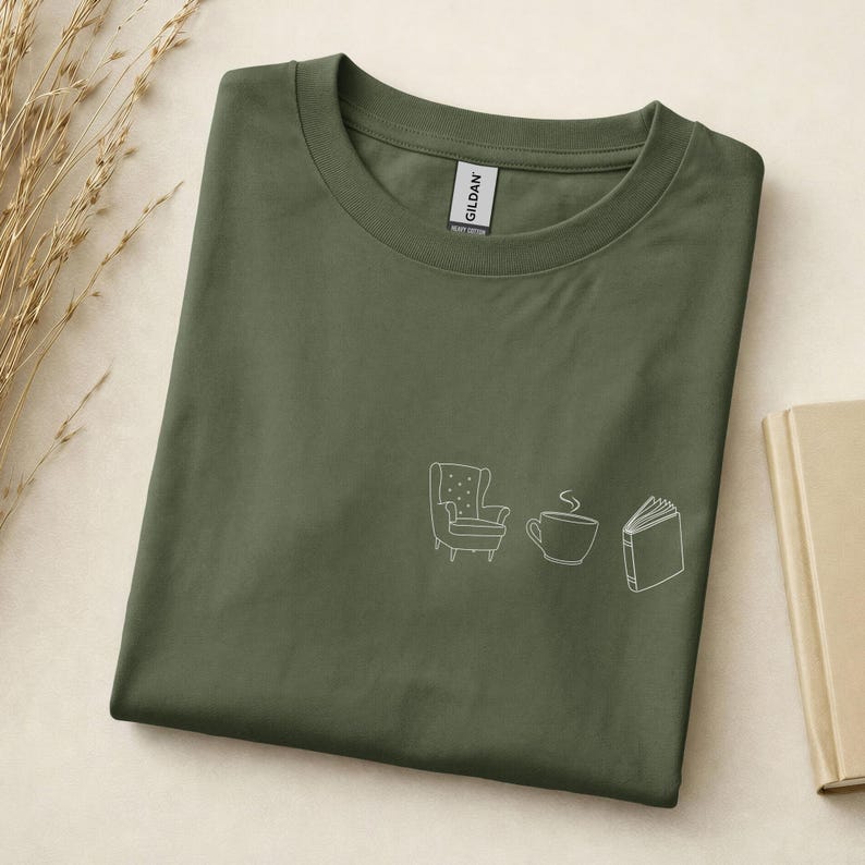 May include: An olive green crew neck t-shirt featuring a white line art graphic on the chest. The graphic depicts a small armchair, a steaming coffee mug, and an open book. The shirt has a Gildan brand label visible at the inner neckline.