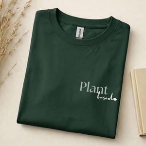 May include: A dark forest green crew neck t-shirt featuring the text Plant based printed in a light sage green and white font on the lower right side. The shirt has a Gildan brand label visible at the inner neckline and a short sleeve design.