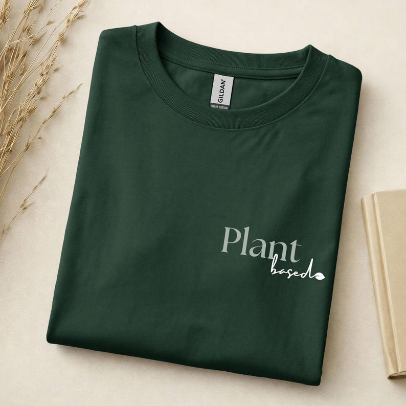May include: A dark forest green crew neck t-shirt featuring the text Plant based printed in a light sage green and white font on the lower right side. The shirt has a Gildan brand label visible at the inner neckline and a short sleeve design.